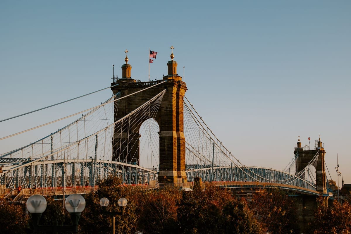 A stone suspension bridge tower with flags on top and cables reaching across a river at sunset