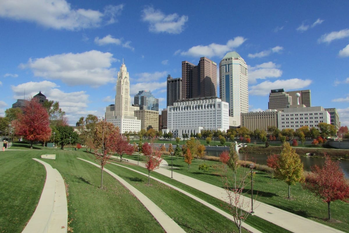 Downtown towers behind a riverside park with curving paths and autumn trees
