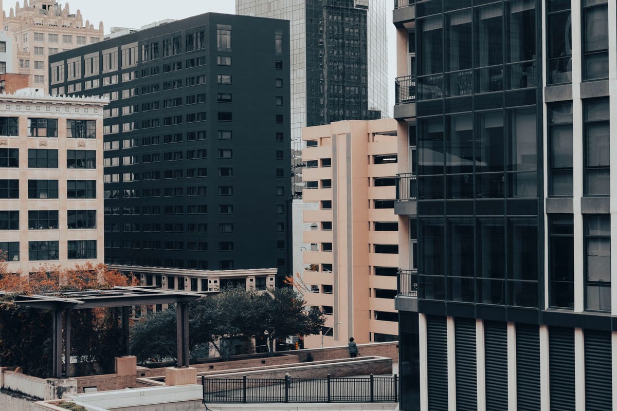Close-set office buildings in black glass, cream stone and brick, with autumn trees and a rooftop walkway below