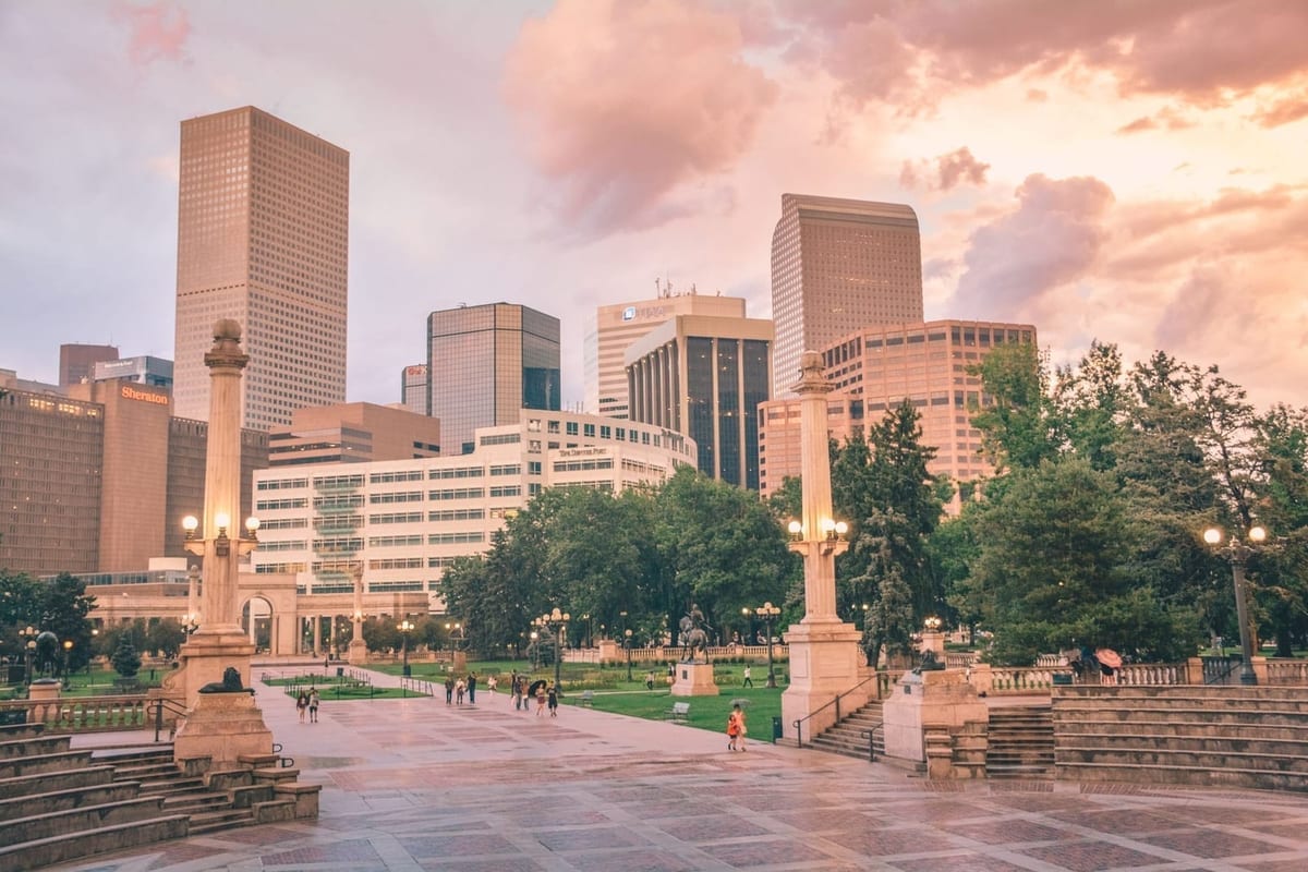 Downtown towers behind a park with paved walkways and lamp posts under a pink sky