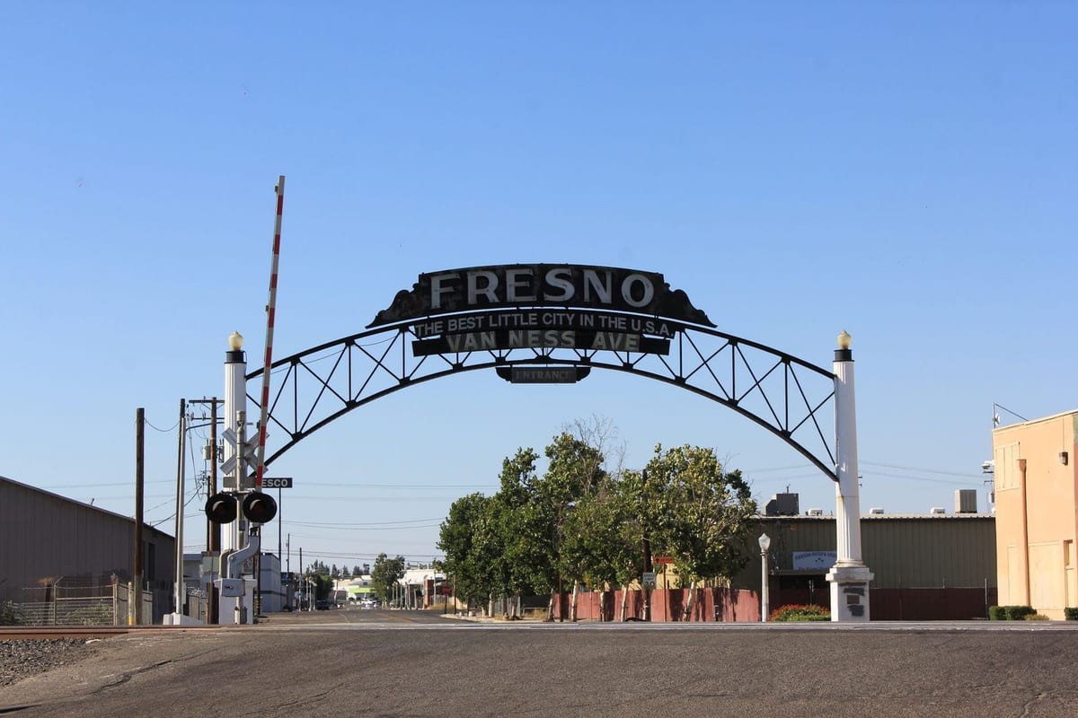 A steel arch with a sign over a railroad crossing, beside a large tree