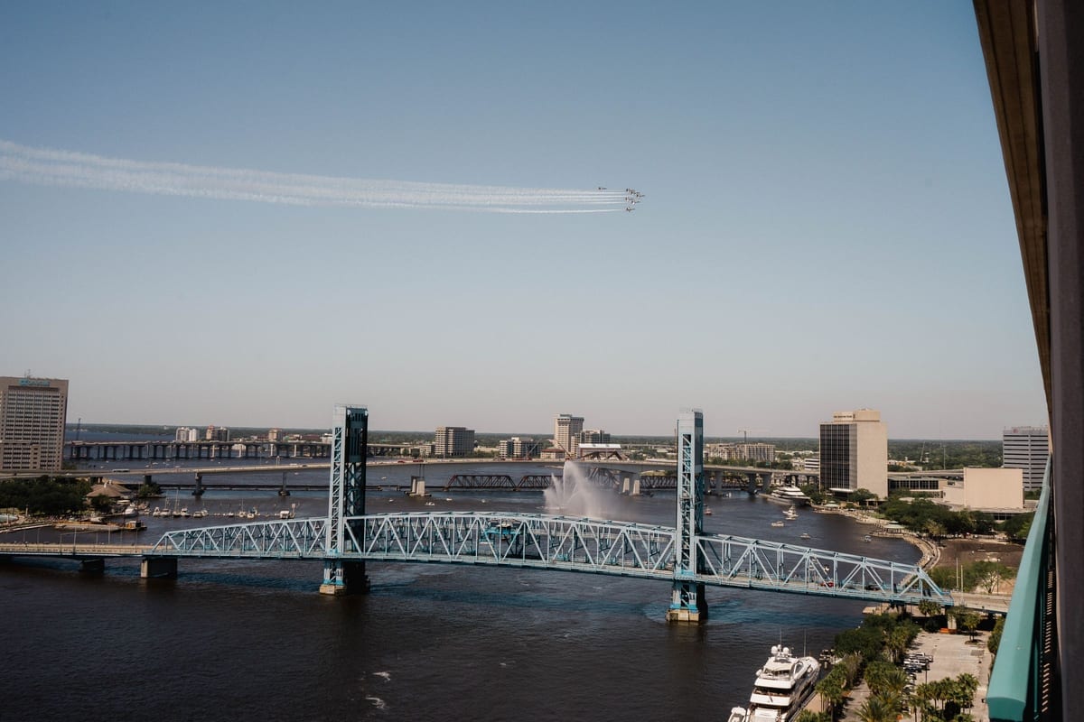 A lift bridge over a wide river beside downtown buildings, with jets trailing overhead