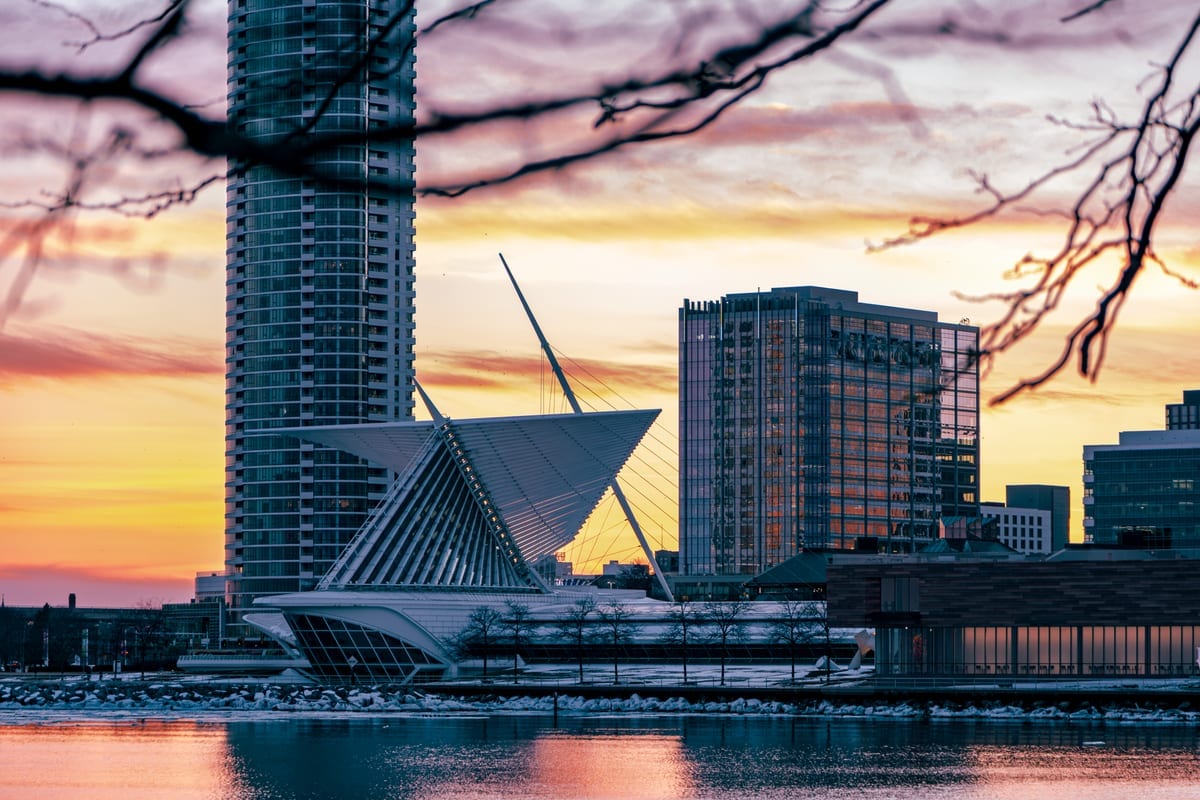 A lakefront building with a winglike roof beside high-rises at sunset, seen through bare branches