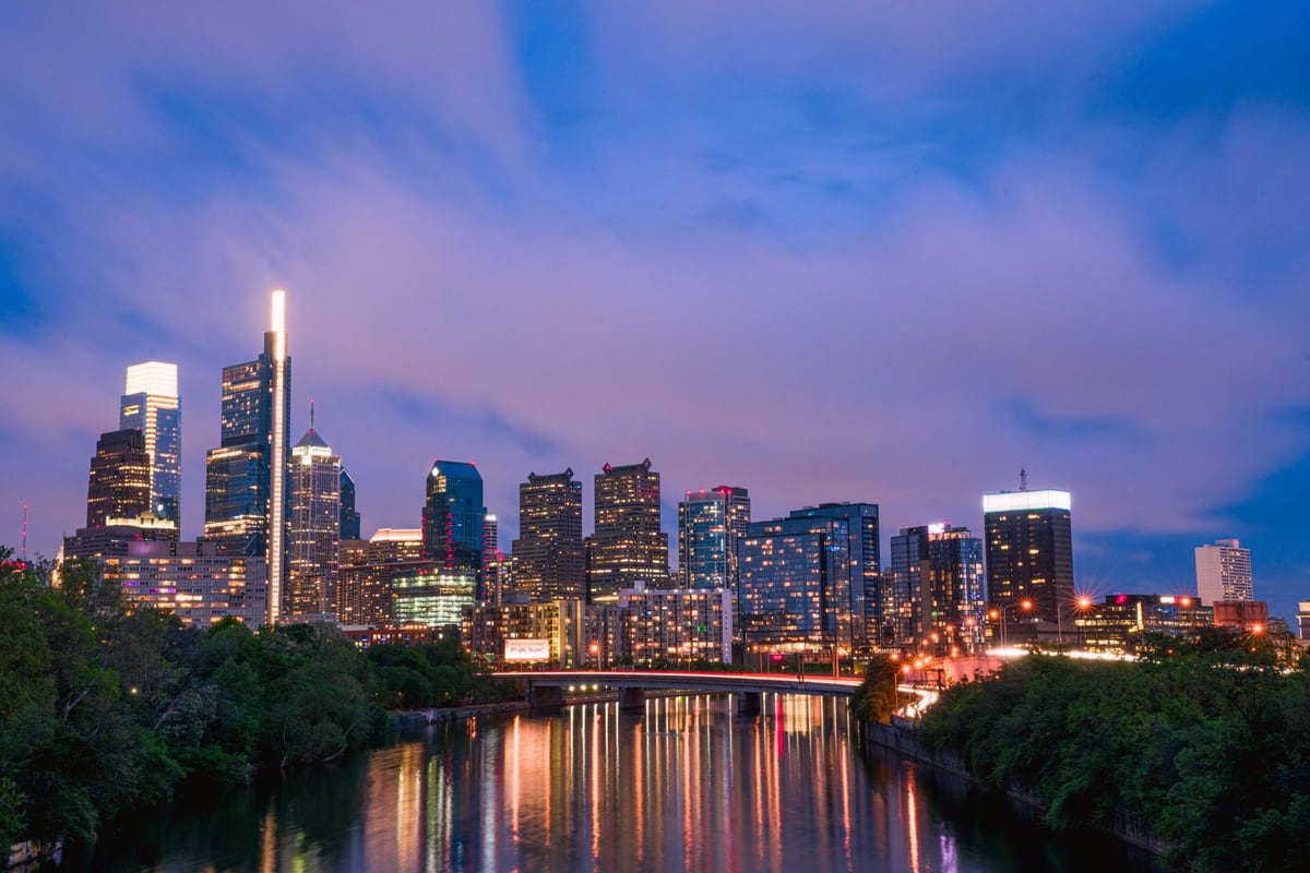 A lit skyline at dusk under purple clouds, reflected in a river with a bridge crossing in front