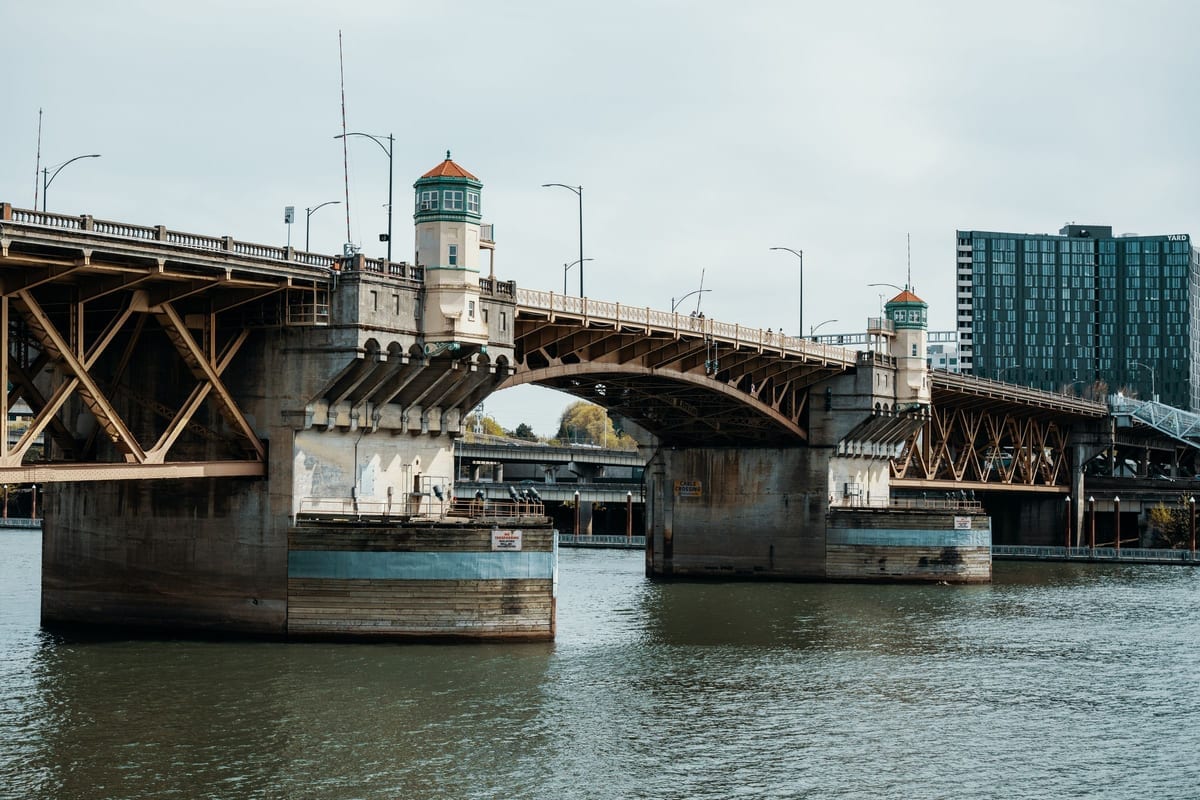 A steel drawbridge with small towers spanning a river, with a glass building beyond