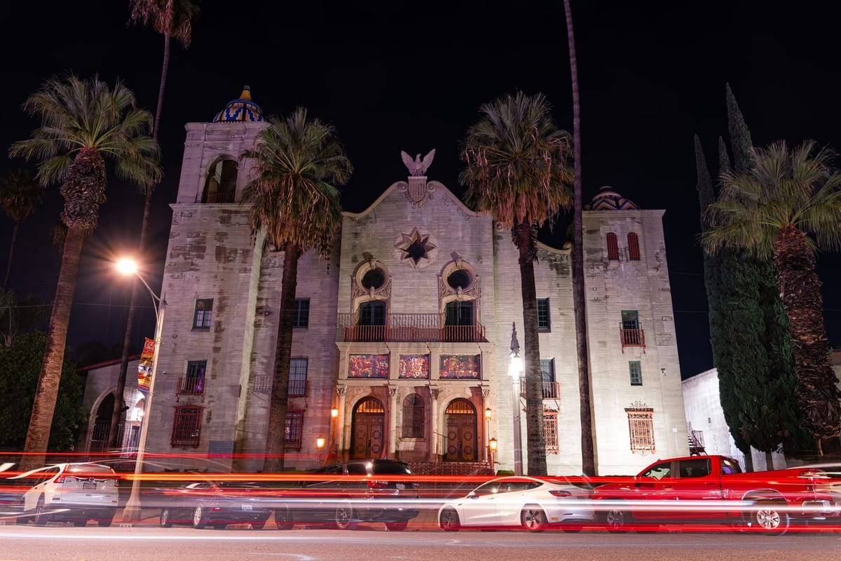 An ornate stone building with bell towers lit at night, with palm trees and light trails from passing cars