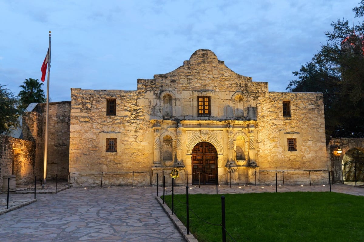 A stone mission facade with an arched doorway, lit at dusk beside a lawn