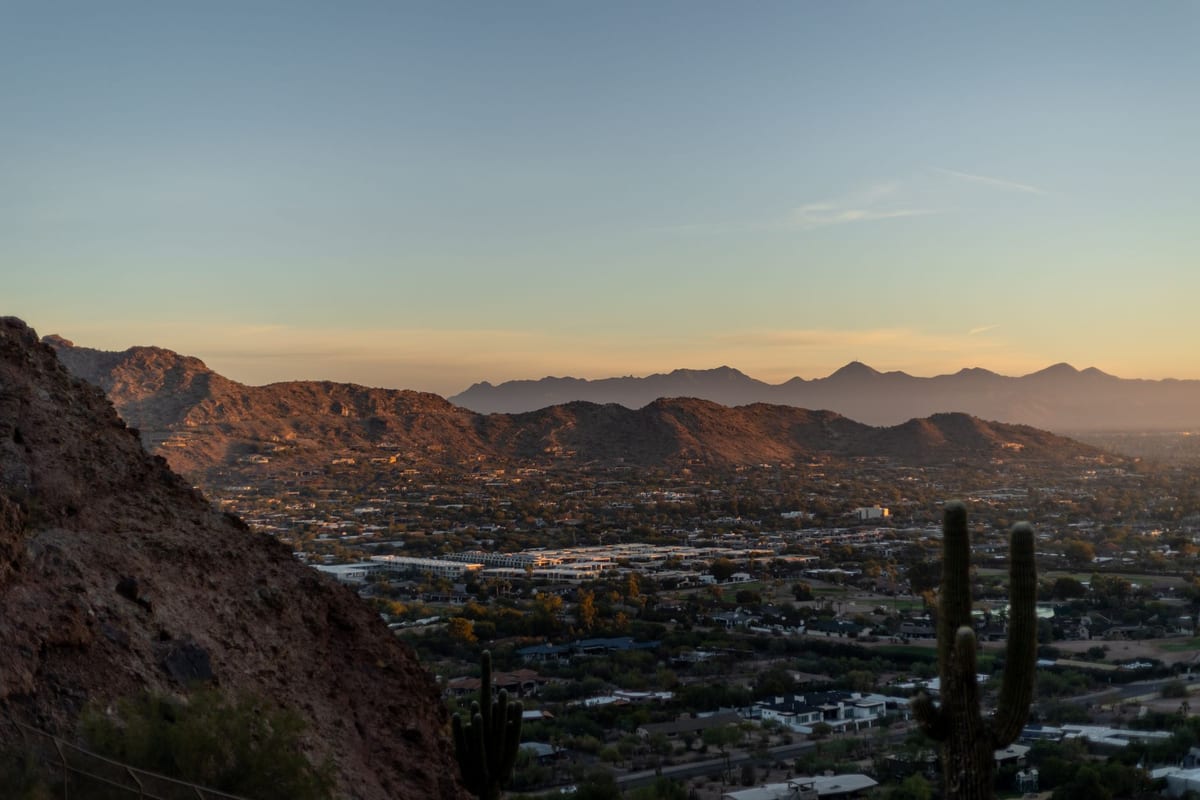 A desert valley city at dusk seen from a rocky hillside with saguaro cactus