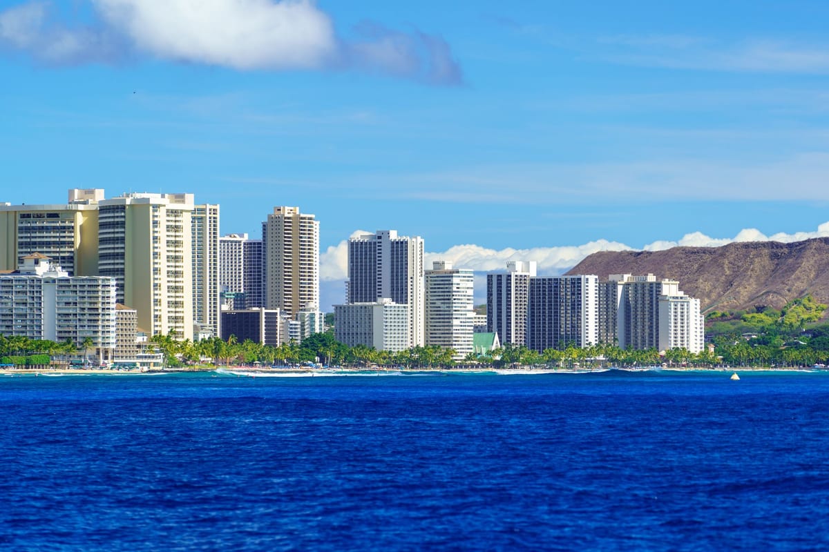 A row of white high-rise towers along a deep blue bay, a green ridge behind them