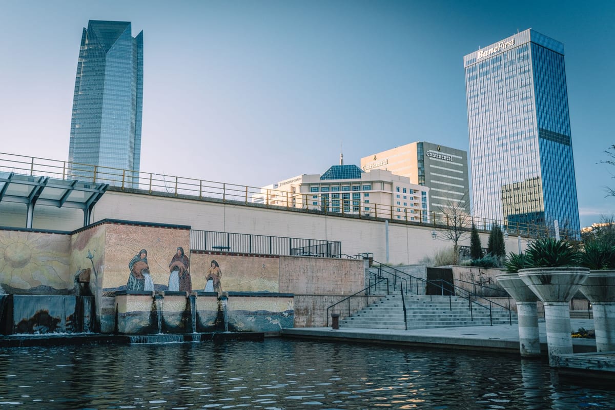 Two glass towers above a concrete riverside walk with sculpted figures along it
