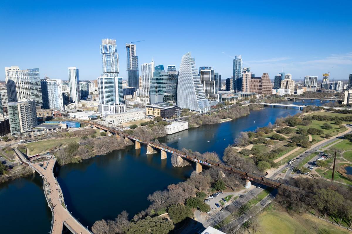 Downtown towers along a river lined with green parkland and a footbridge, seen from above
