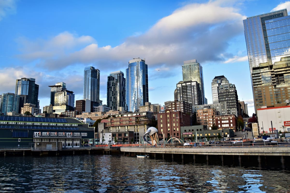 A waterfront skyline of glass towers above piers under blue sky and clouds